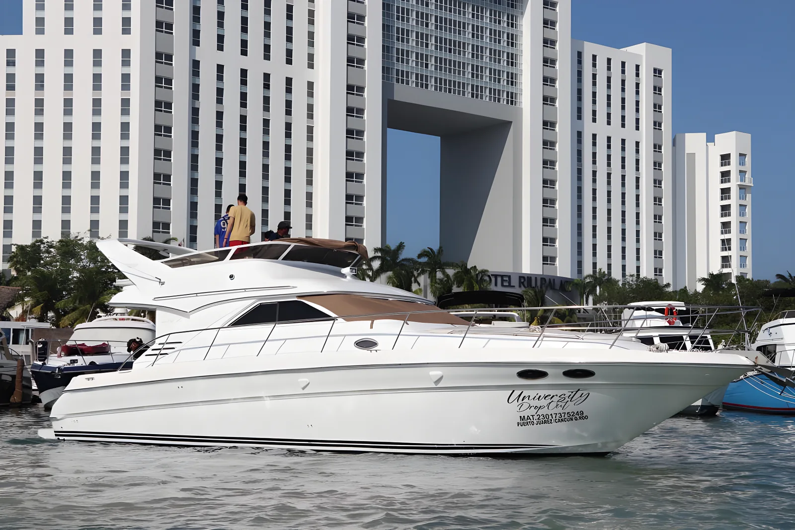 Perfil exterior del yate University Drop Out amarrado con personas en el flybridge y edificios al fondo.